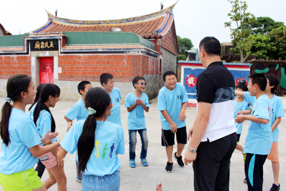 一群穿著淺藍色運動服的小學生在傳統中式建築前的空地上進行戶外體育活動。一位成人教練在中央帶領孩子們進行團體運動或遊戲，背景是具有紅瓦和綠色屋頂的廟宇或傳統建築。
