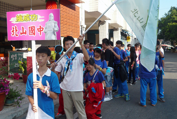 北山國中學校活動，師生穿著藍色制服排隊遊行，手持校旗和標誌牌。校門口掛著粉紅色的學校宣傳看板，周圍有花卉裝飾。隊伍在校園前廣場集合，展現校園活力與團結精神。