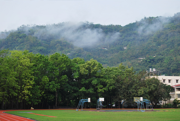 學校運動場景，紅色跑道和綠色草地上有藍色運動器材，遠處是層疊的翠綠山丘，山間籠罩著薄霧，右側可見白色校舍建築，環境優美靜謐。