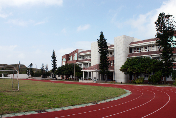 學校校園景觀，紅色跑道環繞綠色草坪，左側可見足球門，背景為紅磚白瓷磚校舍建築，右側種植高大樹木，晴朗天空背景。