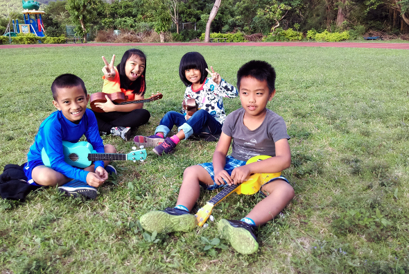 四位兒童坐在公園草地上進行戶外音樂活動，左邊男孩彈著藍色吉他，後方女孩演奏樂器，右邊男孩穿著黃色短褲。背景有綠樹、遊樂設施和紅磚牆。