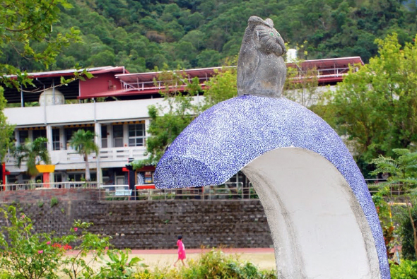 一座藍白相間的大型鯉魚跳躍雕塑矗立在河邊，鯉魚頂部站著一隻鳥。背景為山坡上的建築物和綠色樹木，河邊有人行走。這是一處公共藝術裝置景區。