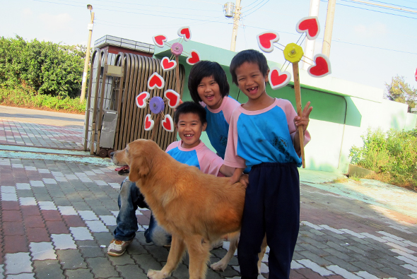 一位成年女性和三個小孩在戶外與一隻棕色狗合照。背景有綠色建築物和心形裝飾品。小孩們穿著藍色和粉紅色衣服，開心地擁抱狗兒。地面鋪著棋盤式磁磚。