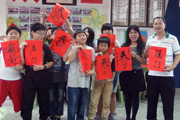 一群學生和成人在教室內舉著紅色春聯，上面寫著中文書法字。背景有黃色和彩色裝飾，牆上貼著照片和海報。眾人面帶笑容，慶祝農曆新年或進行書法文化活動。