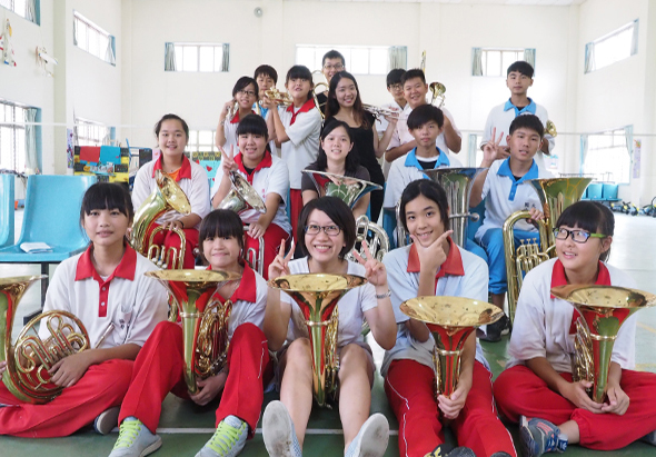 學校音樂社團在室內展示銅管樂器。前排坐著五位學生穿著紅白制服，各持法國號、小號等樂器。後排約十五位學生站立，多數穿著紅白或藍白服裝，手持各式銅管樂器，展示參賽或表演成果。