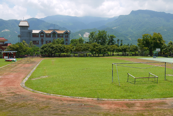 綠色草坪運動場地，設置足球門框，背景為三層樓建築物和翠綠山脈。場地周圍有紅土跑道和樹木，晴朗天空飄浮白雲。