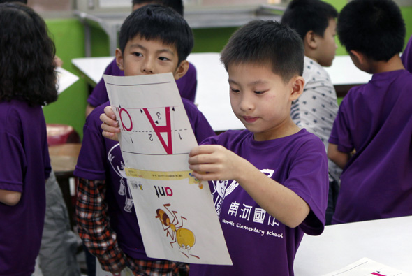 一群穿著紫色制服的小學生在教室內展示自己製作的學習作品。兩名男童在前方舉著繪有數字和圖案的白色紙張，其他同學在周圍觀看，呈現愉快的學習氛圍。