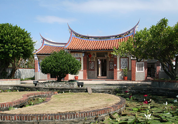 傳統中式廟宇建築，紅磚牆面配橙黃色琉璃瓦屋頂，屋角向上翹起。廟前有荷花池塘，兩側種有綠樹，庭院寬敞。門前放置香爐和供品，展現東方宗教建築特色。