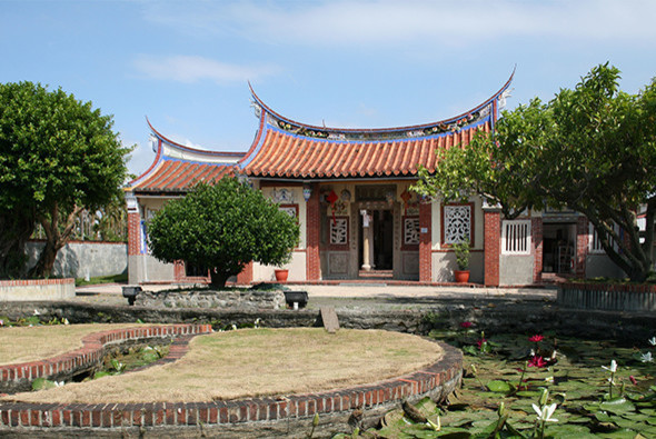 傳統中式廟宇建築，具有紅磚牆體和橙黃色琉璃瓦屋頂，屋頂兩端翹起。前方有荷花池塘，周圍種植綠樹。廟門前方鋪設磚造圓形花壇。建築風格典雅，展現東方傳統宗教建築特色。