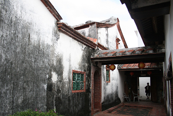 古老的中式建築庭院，白色牆面布滿青苔，木製廊架呈現紅褐色斑駁，綠色木窗框鑲嵌其中，紅磚地面，一人站在內庭門口，展現傳統江南建築風格。
