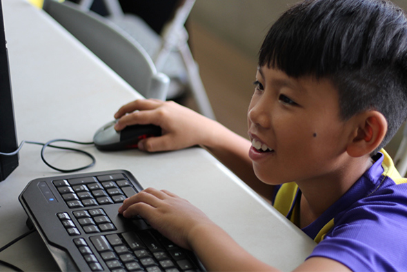 一名穿著紫黃色運動衣的小男孩坐在電腦前，面帶笑容地操作鍵盤和滑鼠，專注於螢幕上的內容。背景可見另一台電腦設備，呈現教室或電腦教學的學習場景。