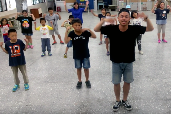 一位年輕男性教練在室內場地帶領約十五名兒童進行團體運動課程。孩子們穿著各色運動服，跟隨教練做出雙臂上舉的動作。背景可見教室環境和其他兒童。這是一堂充滿活力的兒童健身或舞蹈課程。