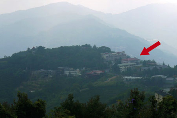 山區景觀，遠處山峰籠罩於霧氣中，山腰處有綠色樹林和散落的建築物，紅色箭頭指向右上方的一棟建築，表示特定地點位置。