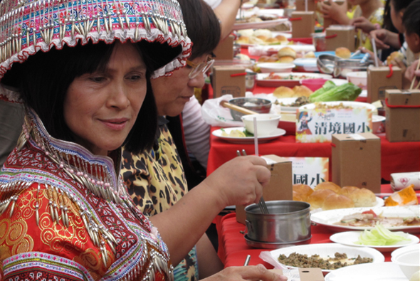 穿著傳統民族服飾的女性在長桌前微笑，手持餐具準備進食。桌上擺放著紅色餐墊、多道菜餚、飲料和食物標籤。背景可見其他參與者和豐盛的餐點，呈現民族文化美食聚餐的場景。