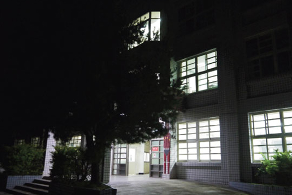 夜間拍攝的建築物外觀，白色瓷磚牆面，多扇大型玻璃窗戶透出溫暖燈光。入口處有紅色標誌牌，周圍種植綠色植物，石板地面，呈現寧靜的夜間景象。