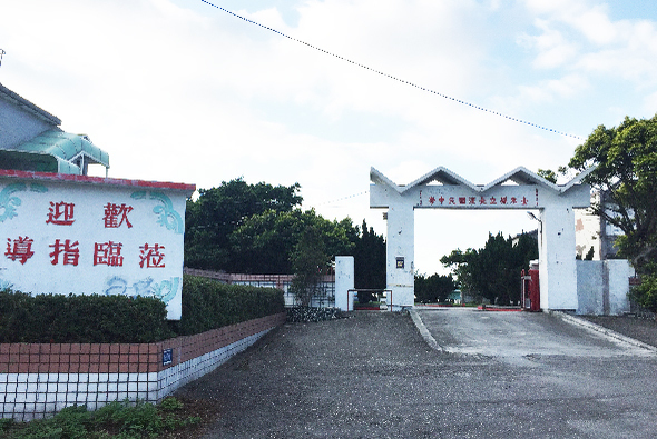 鄉村入口處，左側白牆上書寫紅色歡迎標語，右側為白色牌樓式大門，門楣有中文字樣，道路兩側綠樹環繞，天空晴朗，整體展現典型的中式農村或古鎮景觀。