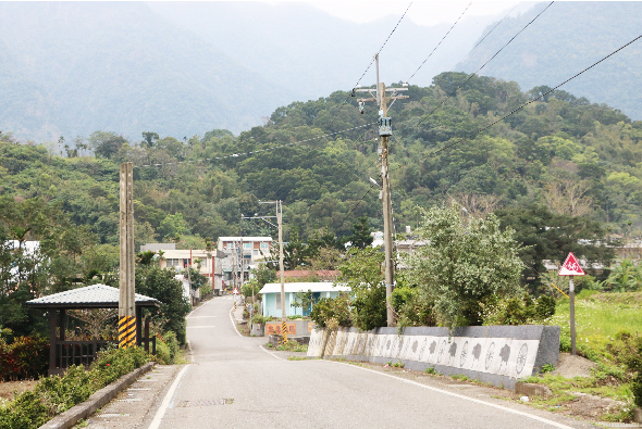 鄉村道路風景，路旁有電線桿、建築物和綠色植被。遠處山丘被茂密森林覆蓋，右側路邊設有白色護欄和警告標誌。天氣晴朗，呈現寧靜的郊區環境。