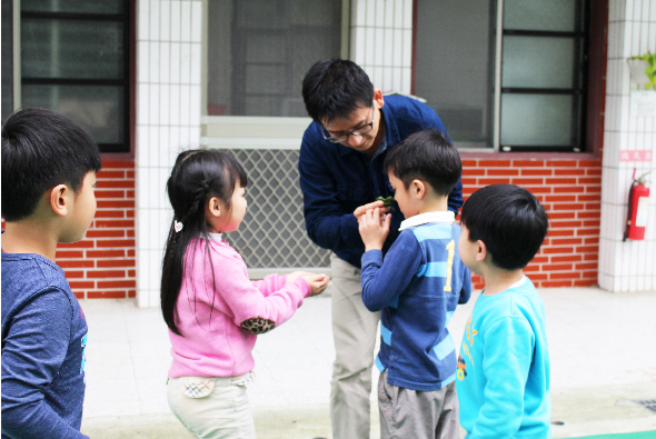 一位成年男性在校園空地上與五位小學生進行互動遊戲。男性穿著深藍色外套，正在指導小孩們進行團體活動。孩子們穿著不同顏色的外套，圍成一圈參與遊戲，背景是紅磚校舍建築。