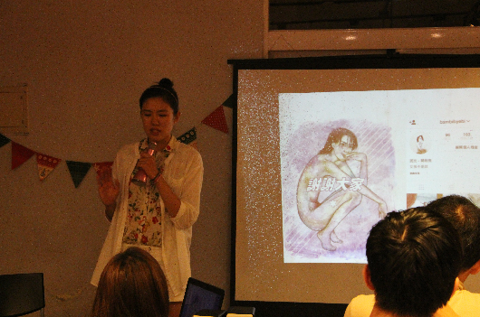 一位女性講者站在投影幕前進行演講，幕上顯示插畫作品。觀眾坐在前方聆聽。會場布置有彩旗裝飾，講者穿著淺色外套，手持麥克風進行介紹。