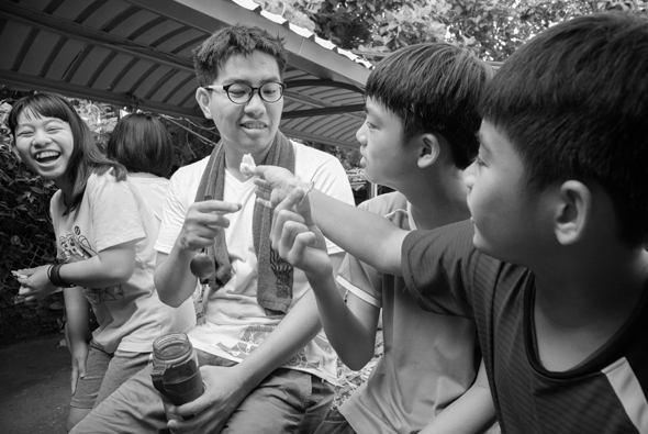 一群人在戶外棚架下進行互動遊戲，中央是戴眼鏡的男性與兩名男孩進行手指遊戲，左側女性和小孩在旁歡樂地觀看，氣氛溫馨愉快。黑白照片呈現親密的家庭或團體活動場景。