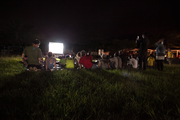 夜間戶外草坪上舉辦露天電影放映活動，眾多大人小孩坐在草地上觀看大螢幕，周圍有帳篷和燈光照明，營造溫馨的社區聚集氛圍。