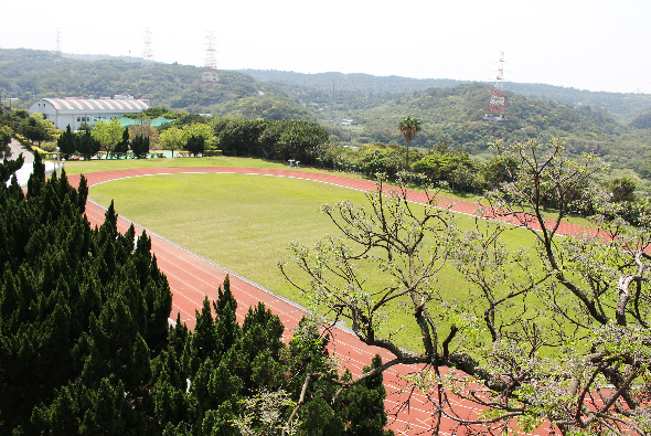 山坡上的運動場景，綠色草坪和紅土跑道環繞，周圍有針葉樹林和建築物。背景為層疊的綠色山脈，展現寧靜的校園或運動設施環境。