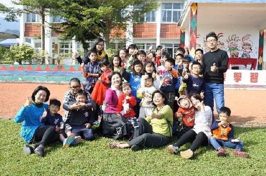 一群小學生和老師在校園草坪上集合拍照。背景是校舍、游泳池和紅色圍牆。學生們穿著各色衣服，有的蹲著，有的站著，展現歡樂的班級合照。
