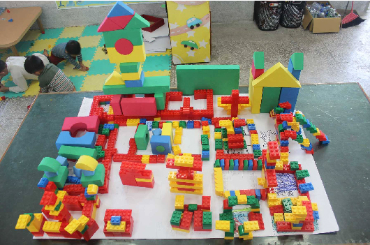 幼兒園教室內，白色地墊上擺放著五顏六色的樂高積木，組成各種建築和圖案。周圍有彩色泡沫地墊、積木牆面裝飾，以及兒童正在進行建構遊戲和創意創作活動的場景。