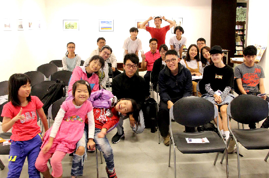 室內活動現場，約二十多位大人和小孩聚集在一起。前排坐著穿著粉紅色衣服的小孩，後排站著青少年和成人。大家面向鏡頭，現場氛圍活潑，牆上掛著圖畫和海報，看起來是社區或教育機構舉辦的集體活動。