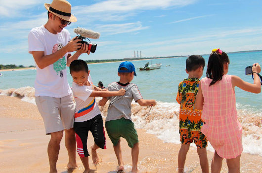 一位成年男性在海灘上用攝影機記錄五個小孩嬉水遊玩的情景。孩子們穿著彩色泳衣，在淺灘邊玩耍，背景是清澈的海水、沙灘和遠處的小船。天空晴朗，陽光普照。