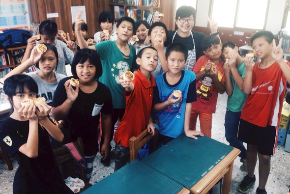 一群小學生在圖書館聚集，圍繞著乒乓球桌，手持食物開心地慶祝。室內有書架、窗戶和室內裝飾，學生穿著各色衣服，氣氛歡樂溫馨。