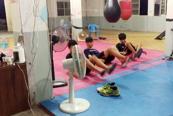 室內運動館中，三名兒童在粉紅色墊子上做伸展運動。地面鋪有藍粉色運動墊，白色風扇放在前方。背景有懸掛的沙袋和健身球，牆上貼有鏡子。兒童穿著運動服參與柔韌性訓練課程。