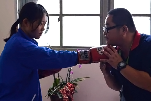 一名穿著藍色衣服的女性拳擊手穿戴紅色拳擊手套，與對面穿著紅色衣服、戴眼鏡的男性進行拳擊練習。背景為窗戶和盆栽植物，室內訓練場景。