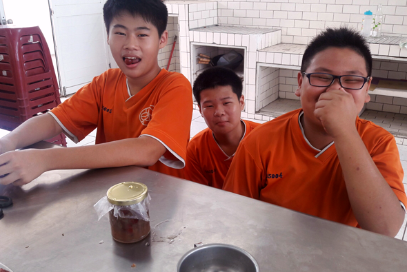 三名穿著橙色制服的年輕人坐在食堂餐桌前，面前放著一罐棕色液體和碗盤。左邊男孩開心地笑著，中間女孩面無表情，右邊戴眼鏡的男孩做出思考狀。背景是廚房設施和紅色籃子。