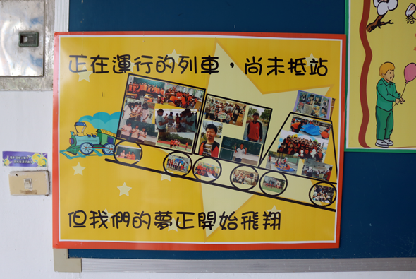黃色海報展示列車造型的活動進度，標題「正在漫行的列車，尚未抵站」，列車車輪內含多張活動照片，下方文字「回我們的夢正開始飛翔」。牆面貼有校園活動宣傳板。