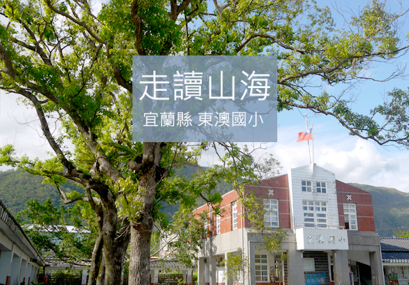 宜蘭縣東澳國小校園，大樹下豎立「走讀山海」白色標誌牌，背景為紅磚校舍建築、山景與飄揚國旗，展現山海環境與在地教育特色。