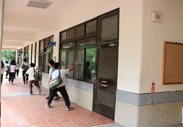 學校走廊內，多名學生穿著運動服進行體能訓練。他們在寬敞的紅磚地面上做出各種動作，如跳躍和伸展。走廊兩側有大型玻璃窗和教室門，天花板裝有照明設備，整個空間明亮寬敞。