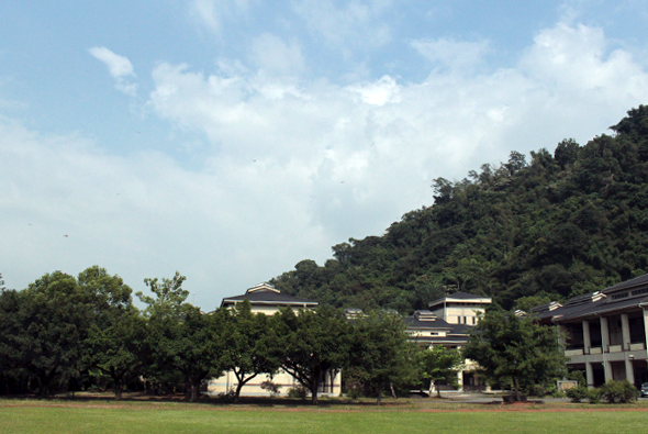寬闊的綠色草坪前方，矗立著數棟古典風格的黃色建築，右側為殖民地風格的廊柱建築。左邊高大的樹木提供遮蔭，背景為茂密的山林，藍天白雲點綴，呈現歷史校園或官方機構的典雅景觀。