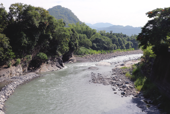 寬闊的河流流經山谷中央，河水呈現灰綠色，兩側布滿石礫。河岸邊生長著茂密的樹木和植被，背景是層疊的綠色山脈和遠方的山峰，形成壯闊的山水風景。