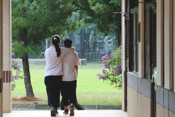 兩位女性並肩站在建築物走廊的出入口，面向室外的草坪和樹木。走廊兩側有紫色花卉盆栽，陽光灑落在綠色草地上，呈現寧靜的校園或機構環境。