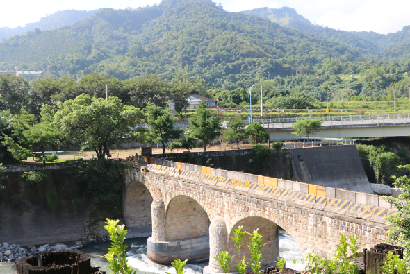 古色古香的石拱橋跨越河流，橋面鋪有黃色施工標示帶。背景為層疊青山與茂密樹林，前景可見河床與綠色植被。右方有現代混凝土水利設施，形成古今對比的風景。