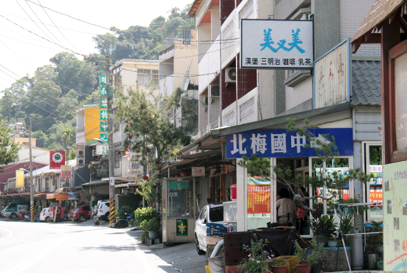 台灣山區街道景觀，兩側為多層樓房和傳統店鋪。右方可見「北梅國中」和綠色招牌店面，左方有黃色廣告牌和紅色機車。山坡與電線杆點綴背景，呈現典型鄉村商業區的風貌。