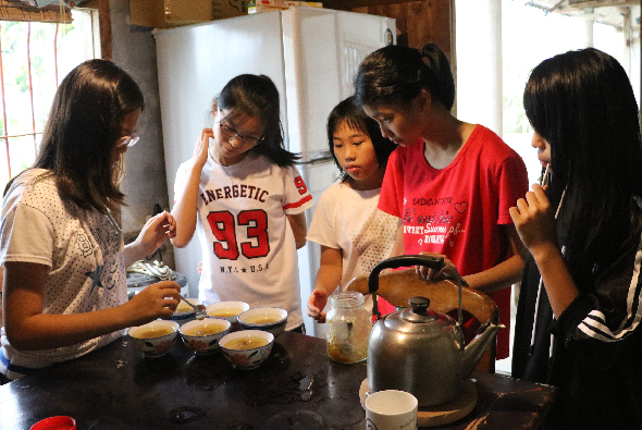 五位年輕女性在廚房圍著料理台，聚精會神地準備食物。其中一人穿著紅色T恤正倒水入茶壺，桌上放著碗盤和食材。她們正進行烹飪或料理教學活動。