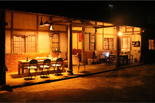 夜間照亮的建築物，展示傳統磚造房舍的室內外空間。左側為用餐區，擺放餐桌和椅子；右側為辦公或工作區域，放置電腦設備。溫暖的黃色燈光照亮整個空間，營造溫馨的夜間氛圍。