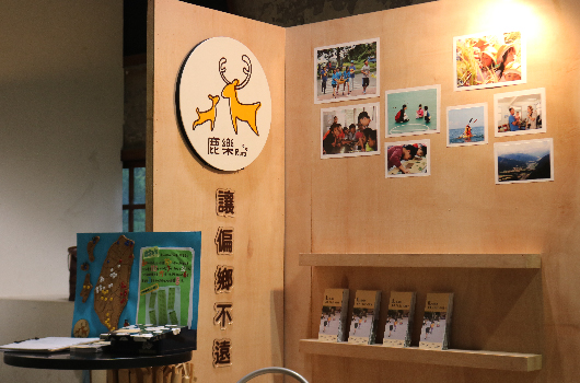 展覽區展示鹿茸相關產品和文化。牆面貼有鹿標誌和中文文字，右側陳列家庭照片，左側放置茶具和竹林圖片，下方展示鹿茸產品包裝盒。整體呈現溫暖的木質調展示空間。