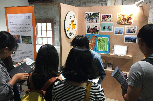 四位參展者在室內展覽空間觀看牆上展示的照片和藝術作品。左側有橙色的說明看板，牆面貼滿各式相片、手繪圖案及彩色裝飾框，展現創意展覽內容。