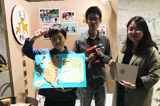 四位學生在教室內展示專題作品。左側學生舉著繪有地圖的藍色展板，中間兩位學生分別持有製作工具和作品，右側學生手持資料夾。牆面後方貼有多張照片和學習成果。