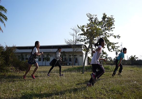 五名學生在校園空地上進行戶外運動。背景為現代校舍建築，周圍種有樹木。學生們穿著運動服裝，在草地上跑步和活動，享受陽光充足的戶外體育課程。