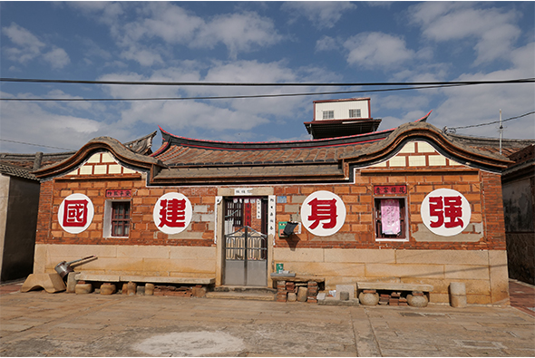 一棟紅磚平房建築，正面牆上裝飾著四個白色圓形圖案，內有紅色繁體中文字。屋頂為傳統翹角設計，上方放置現代水塔。門前空地簡樸，電線橫跨上空。建築展現傳統與現代混合的風格。