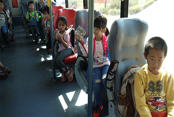 公車車廂內，多名小學生坐在紅色和灰色座椅上。右側穿黃色上衣的男童看向窗外，中間女童穿著粉紅色外套，左側有其他學童。陽光透過車窗灑入車廂，顯示這是一次學校集體搭乘公車的校外教學活動。